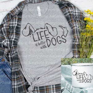 May include: A gray t-shirt with the text "Life is better with dogs" and a dog silhouette. The shirt is displayed on a plaid shirt and a wooden surface. A white enamel mug with the same design is also shown.