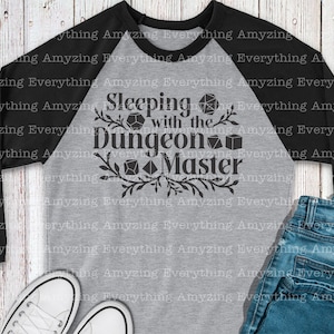 May include: A gray and black baseball tee shirt with the text "Sleeping with the Dungeon Master" in black. The text is surrounded by a black floral design and black dice.