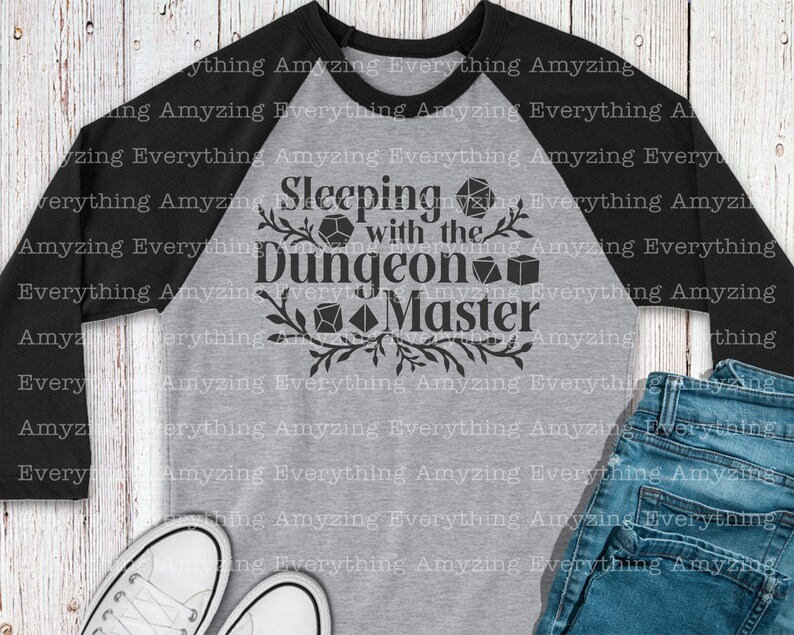 May include: A gray and black baseball tee shirt with the text "Sleeping with the Dungeon Master" in black. The text is surrounded by a black floral design and black dice.