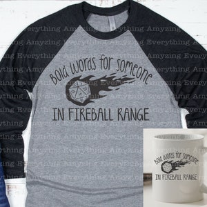 May include: A gray and black baseball tee shirt with a black graphic of a 20-sided die on fire with the text "Bold words for someone in fireball range". A white mug with the same graphic is shown to the right.