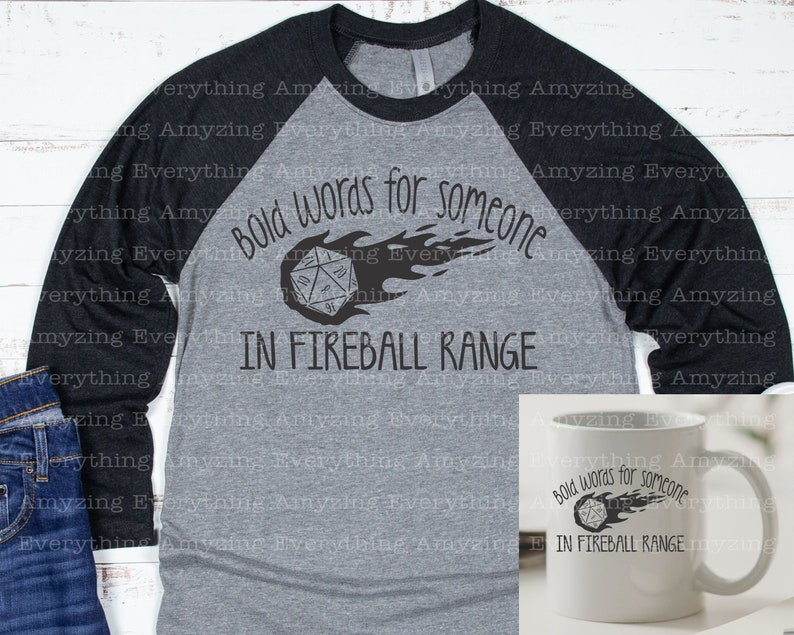 May include: A gray and black baseball tee shirt with a black graphic of a 20-sided die on fire with the text "Bold words for someone in fireball range". A white mug with the same graphic is shown to the right.