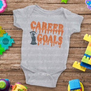 Puede incluir: Un body de beb&eacute; gris con el texto "CAREER GOALS" en letras naranjas que gotean. Una silueta negra de la Muerte est&aacute; a la izquierda del texto.