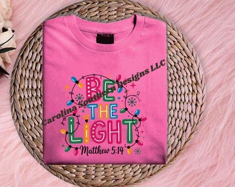 Peut inclure: Un t-shirt rose avec les mots "BE THE LIGHT" en lettres colorées et une guirlande de lumières de Noël. Le texte "Matthew 5:14" est en dessous. Le t-shirt est sur un dessous de plat tressé.