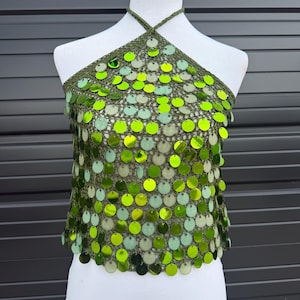 May include: A green halter top with a crochet design and large, round, green sequins. The top is displayed on a white mannequin. The sequins are in various shades of green, creating a shimmering effect.