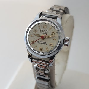 May include: A silver wristwatch with a white face and gold accents. The watch face has black Roman numeral hour markers and black hands. The watch band is a silver metal bracelet. The watch face says "Calvan 17 Jewels Incabloc Waterproof Antimagnetic".