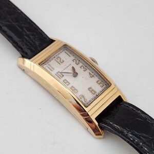 May include: A gold-toned Waltham wristwatch with a white face and black leather band. The watch has a rectangular face with black Roman numeral hour markers and black Arabic numeral minute markers.