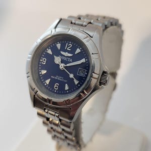 Peut inclure: Une montre Invicta en argent avec un cadran bleu et un bracelet en métal argenté. La montre a un guichet date à 3 heures et les mots "Quartz Water Resistant" imprimés sur le cadran.