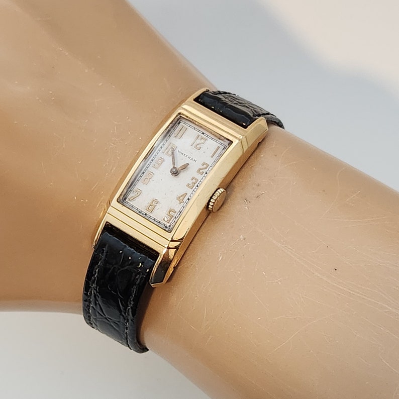 May include: A gold-toned wristwatch with a rectangular face and black leather band. The watch face has Roman numeral hour markers and the word "Waltham" printed on it.