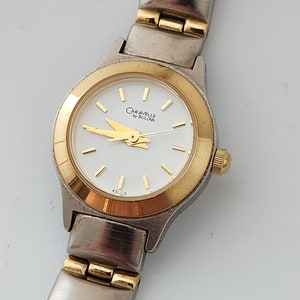May include: A two-tone gold and silver women's wristwatch with a white face. The watch face has gold hands and markers and the text "Caravelle by Bulova" printed in gold. The watch has a gold bezel and a two-tone band.