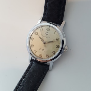 Peut inclure: Une montre-bracelet argentée avec un bracelet en cuir noir. Le cadran de la montre est de couleur crème avec des chiffres et des aiguilles noirs. Le cadran de la montre porte également le texte "Medallist" et "17 Jewels Incabloc".