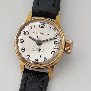 May include: A gold-tone wristwatch with a white face and black leather strap. The watch face has black hour and minute hands, a black second hand, and black numbers. The watch face also has the text "DUMAI" and "21 JEWELS INCABLOC".