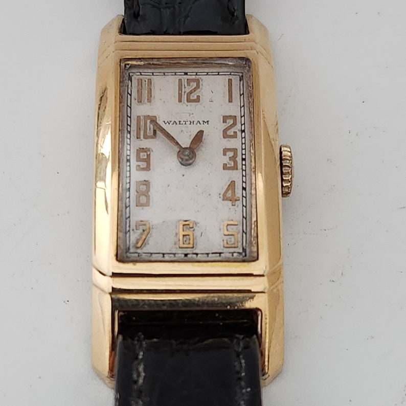 May include: A gold-toned wristwatch with a white face and black leather band. The watch face has black Roman numeral hour markers and a 'Waltham' logo.