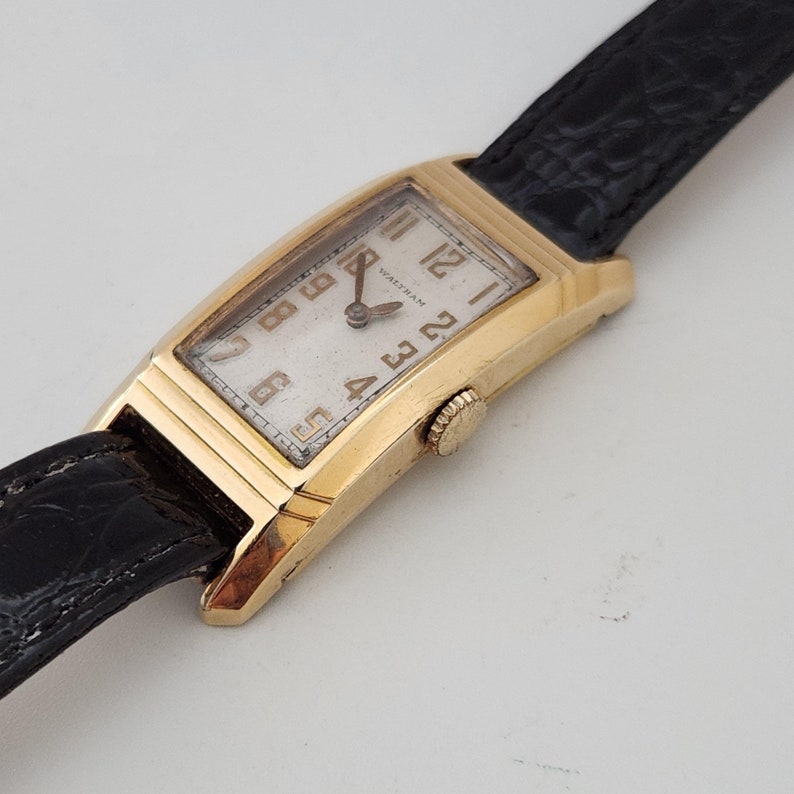 May include: A gold-toned Waltham wristwatch with a rectangular face and black leather band. The watch face has Arabic numerals and a white background.