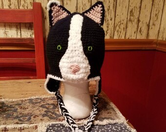 Puede incluir: Un gorro de punto negro y blanco con forma de gato, con ojos verdes y nariz rosa. El gorro tiene un cordón largo trenzado con borlas.