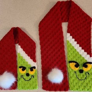 May include: Two crocheted scarves featuring a Grinch design. The scarves are red and green with white accents. The smaller scarf has a white pom-pom on the end. The larger scarf has a white pom-pom on one end and a green Grinch face on the other end.