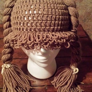 Puede incluir: Un gorro de punto marrón con dos trenzas marrones largas. Las trenzas tienen detalles de hilo blanco en los extremos. El gorro está en una cabeza de maniquí blanca.