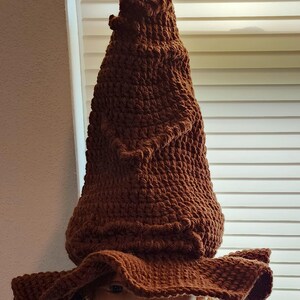 May include: A brown, crocheted hat with a tall, pointed crown and a wide, floppy brim. The hat is made of a textured yarn, and the color is a warm, earthy brown. The hat is displayed on a mannequin head.