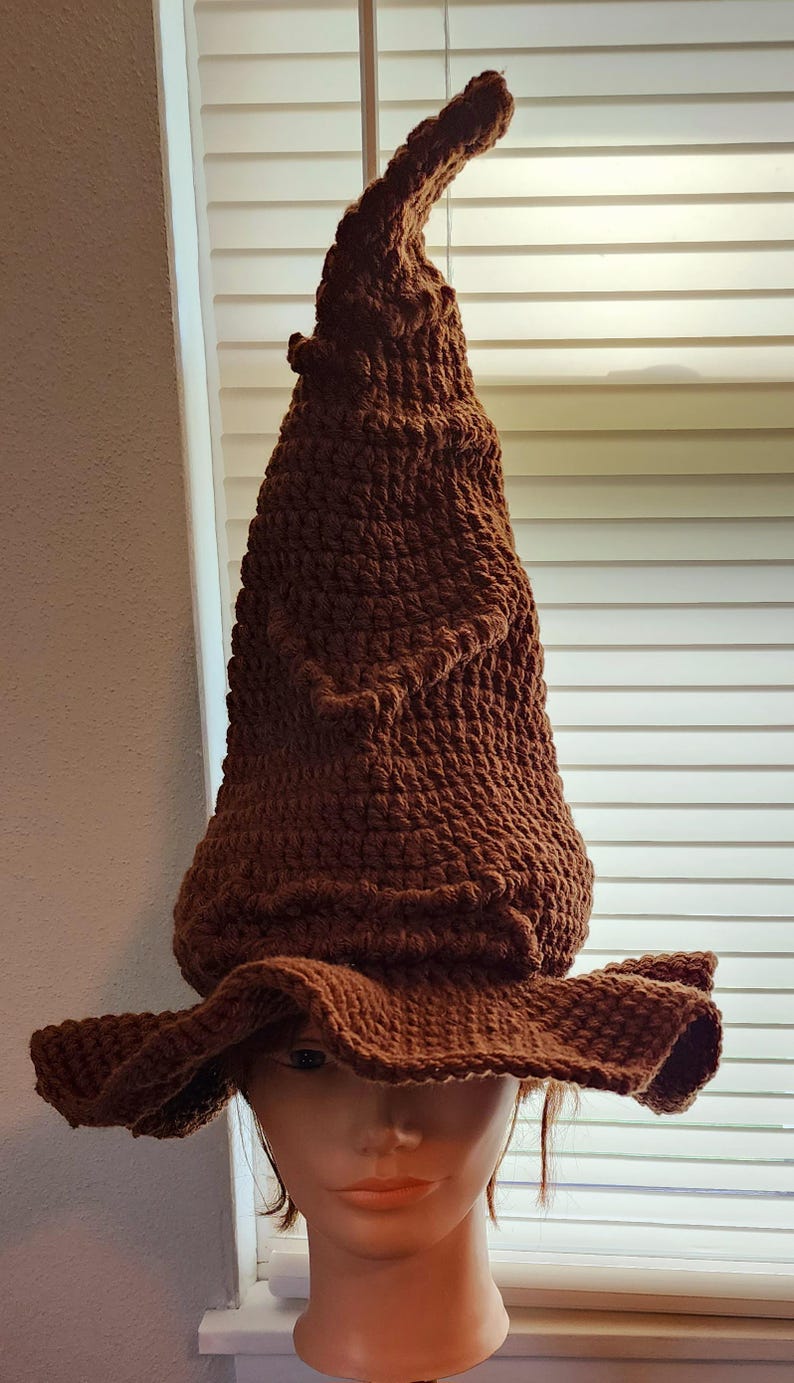 May include: A brown, crocheted hat with a tall, pointed crown and a wide, floppy brim. The hat is made of a textured yarn, and the color is a warm, earthy brown. The hat is displayed on a mannequin head.