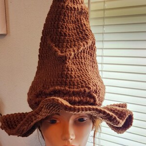 May include: A brown, handmade crochet wizard hat with a pointed crown and wide brim. The hat is displayed on a mannequin head, showcasing its textured stitch and shape. The hat is a costume accessory.