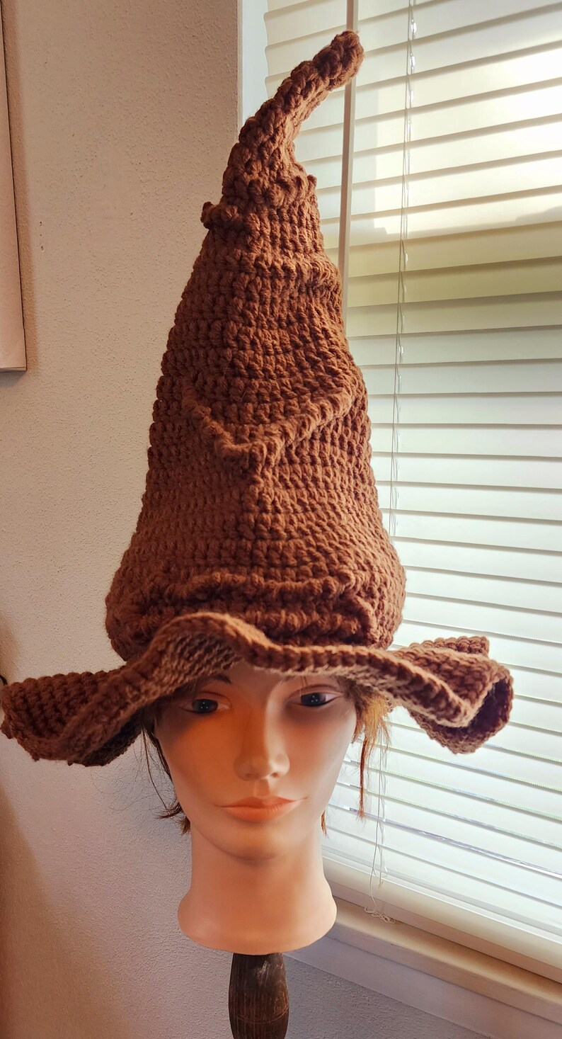 May include: A brown, handmade crochet wizard hat with a pointed crown and wide brim. The hat is displayed on a mannequin head, showcasing its textured stitch and shape. The hat is a costume accessory.