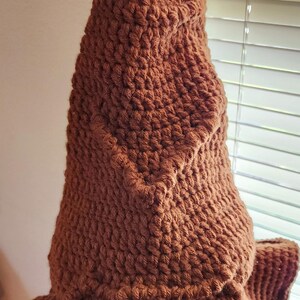 May include: A brown, handmade crochet hat with a pointed top and a wide brim. The hat is made of a textured yarn, and the color is a warm, earthy brown. The hat is likely a costume or novelty item.