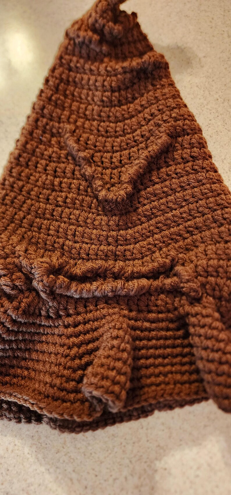 May include: Close-up of a brown crocheted hat with a textured surface. The hat features a detailed, raised design, possibly a tree or leaf pattern, adding a unique touch to the handmade item.