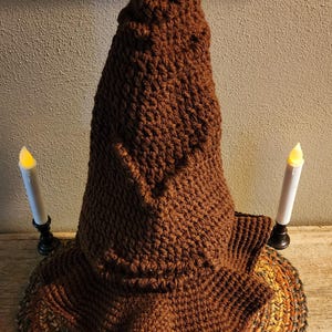 May include: A brown, handmade crochet wizard hat with a wide brim. The hat is displayed with two battery-operated candles on a wooden surface. The hat's texture and color suggest a cozy, handcrafted item, perfect for costume or decor.