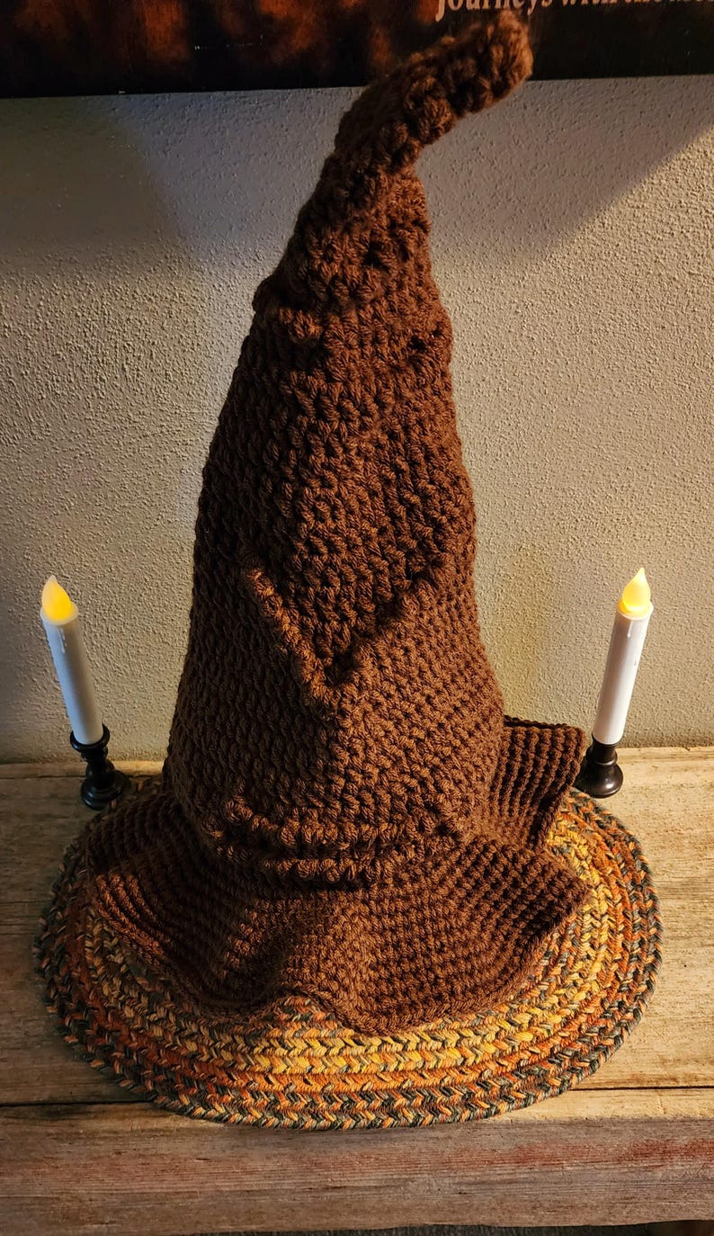 May include: A brown, handmade crochet wizard hat with a wide brim. The hat is displayed with two battery-operated candles on a wooden surface. The hat's texture and color suggest a cozy, handcrafted item, perfect for costume or decor.