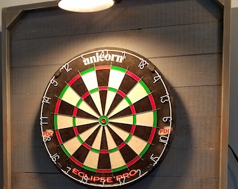 Puede incluir: Un tablero de dardos negro y verde con el texto "unicorn eclipse pro" y "PDC" en él. El tablero está montado en una pared de madera con una luz encima. Tres dardos están clavados en el tablero.