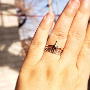 Könnte beinhalten: Ein silberner Ring mit einem Knotendesign und einem Kreuzanhänger. Der Ring wird am Ringfinger einer Hand getragen.