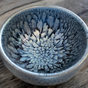 Puede incluir: Un cuenco pequeño y redondo de cerámica con un diseño interior texturizado en azul y blanco. El cuenco tiene un borde plateado y un acabado brillante. El patrón interior se asemeja a un diseño floral o de estrella. El cuenco se utiliza probablemente para servir alimentos o como objeto decorativo.