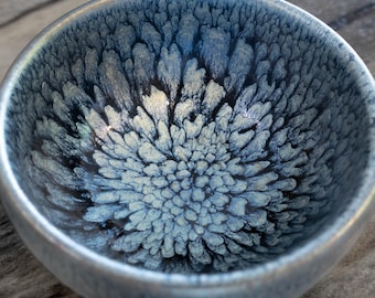 May include: A small, round ceramic bowl with a textured, blue and white interior design. The bowl has a silver-colored rim and a glossy finish. The interior pattern resembles a floral or starburst design. The bowl is likely used for serving food or as a decorative item.