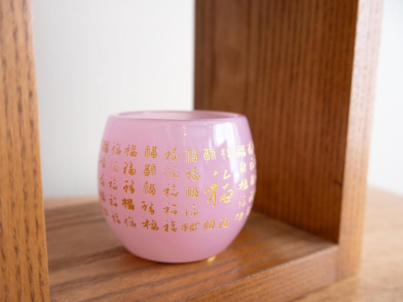 May include: A pink glass candle holder with gold Chinese characters. The round holder sits on a wooden shelf. The candle holder has a soft, translucent appearance.