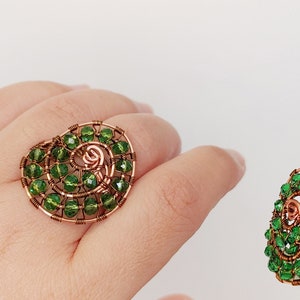 May include: A handcrafted copper wire ring featuring a spiral design, embellished with green beads. The ring is shown on a hand, highlighting its unique, artistic style. Additional views of the ring are shown on a white background.