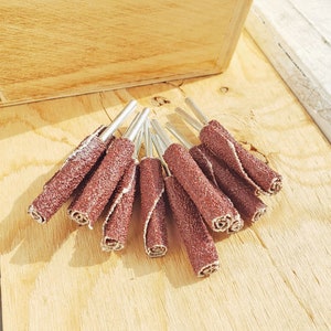 May include: A close-up of a bundle of brown sanding drums with silver metal shafts. The sanding drums are used for sanding and polishing wood, metal, and other materials.
