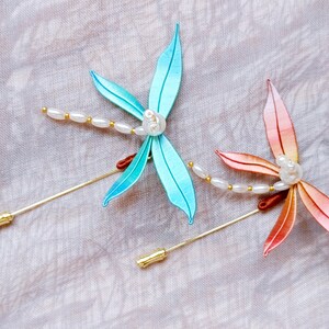 May include: Two delicate dragonfly brooches with pearl accents. One is a light blue colour and the other is a peachy pink colour.