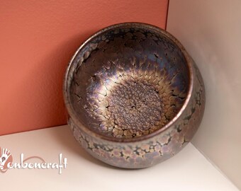 May include: A handcrafted, iridescent ceramic bowl with a textured interior. The bowl's exterior has a dark, speckled finish, while the inside features a unique, crackled pattern with shades of gold and purple. The bowl is set against a peach-colored background.