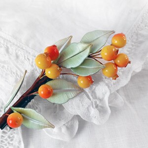May include: A brown twig with green leaves and orange and yellow berries. The twig is a hair clip.