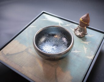 May include: A black ceramic teacup with a silver glaze sits on a tray with a blue and white design. A small ceramic figurine is also on the tray.