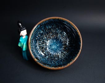 May include: A dark blue ceramic teacup with a white and brown glaze. The glaze has a swirling pattern that resembles a galaxy.