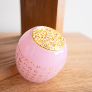 May include: A pink, spherical decorative object with gold-colored accents. The top features an ornate gold design, and gold characters are printed on the pink surface. The object is resting on a wooden surface.
