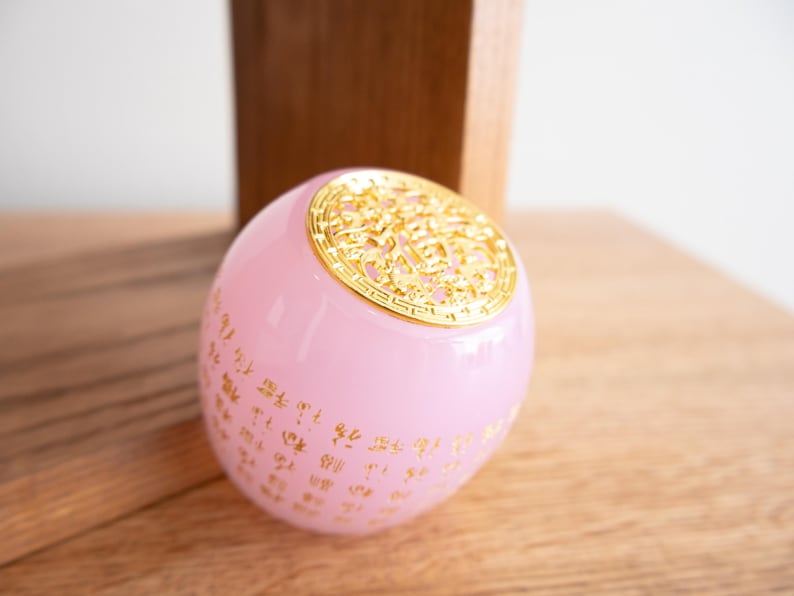 May include: A pink, spherical decorative object with gold-colored accents. The top features an ornate gold design, and gold characters are printed on the pink surface. The object is resting on a wooden surface.