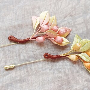 Può includere: Due delicati fermagli floreali con spille dorate e steli marroni. Uno presenta fiori rosa e bianchi, mentre l'altro ha fiori gialli e arancioni.