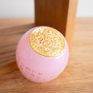 May include: A pink, spherical decorative object with gold-colored filigree on top and gold script around the base. The object is sitting on a wooden surface.
