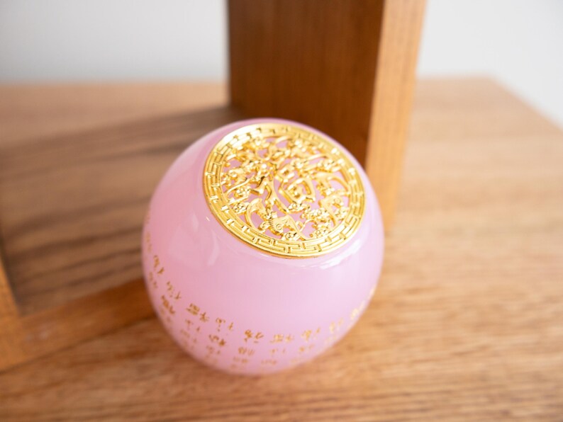 May include: A pink, spherical decorative object with gold-colored filigree on top and gold script around the base. The object is sitting on a wooden surface.
