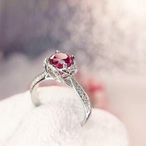 May include: A silver ring featuring a large, round, red gemstone. The ring's band is set with small, clear gemstones. The setting around the red stone has a floral design.