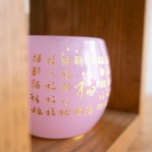 May include: A light pink glass bowl with gold Chinese characters. The bowl is inside a wooden shelf. The bowl has a rounded shape and a smooth surface. The gold characters are evenly spaced around the bowl.