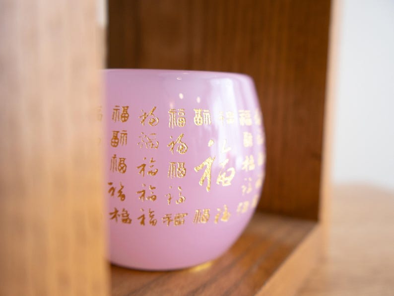May include: A light pink glass bowl with gold Chinese characters. The bowl is inside a wooden shelf. The bowl has a rounded shape and a smooth surface. The gold characters are evenly spaced around the bowl.
