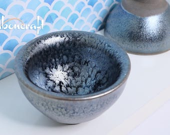 May include: A small, round ceramic bowl with a textured, dark blue glaze and a lighter, white interior. The bowl is part of a set, with a matching piece visible in the background. The background features a blue and white pattern.