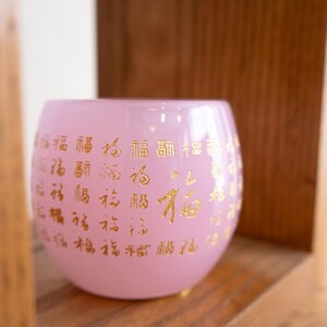 May include: A pink glass votive candle holder with gold Chinese characters. The round candle holder sits on a wooden shelf. The candle holder is a soft pink color.